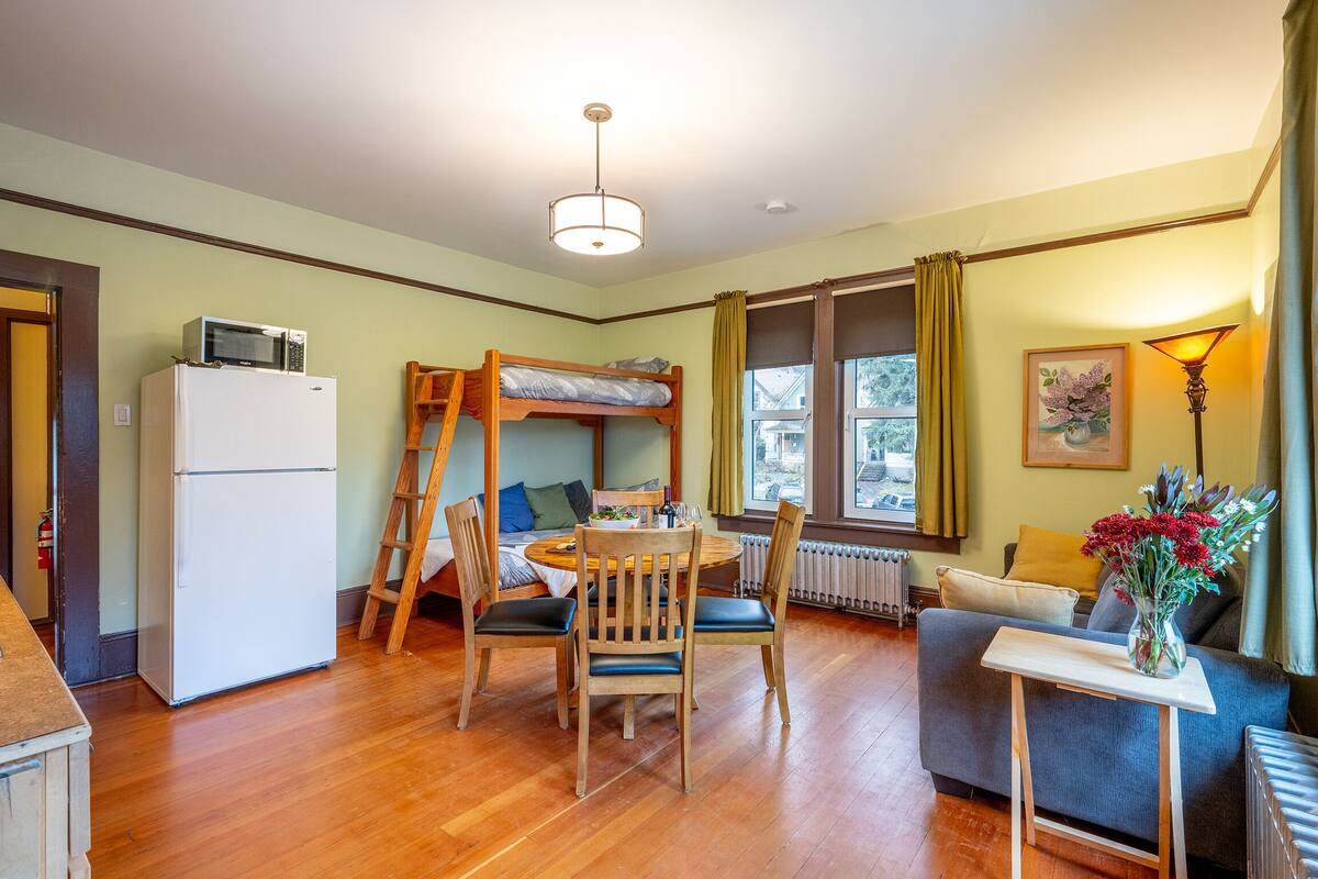 Ein kompakter Wohnraum mit Etagenbett, kleinem Kühlschrank, Esstisch mit Stühlen, Sofa und Lampe.