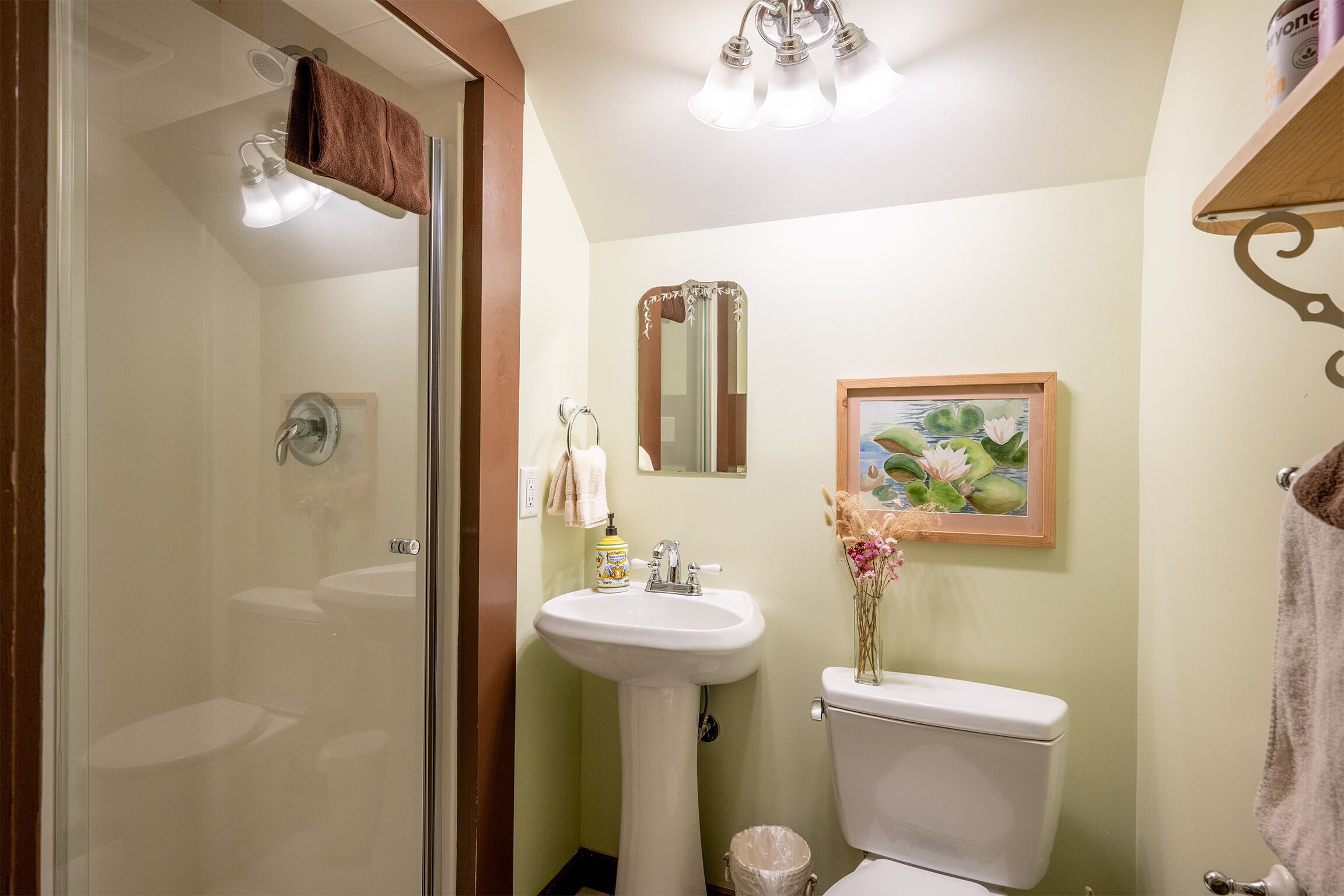 A bathroom with a glass shower enclosure, a pedestal sink, a toilet, and a framed picture of water lilies.