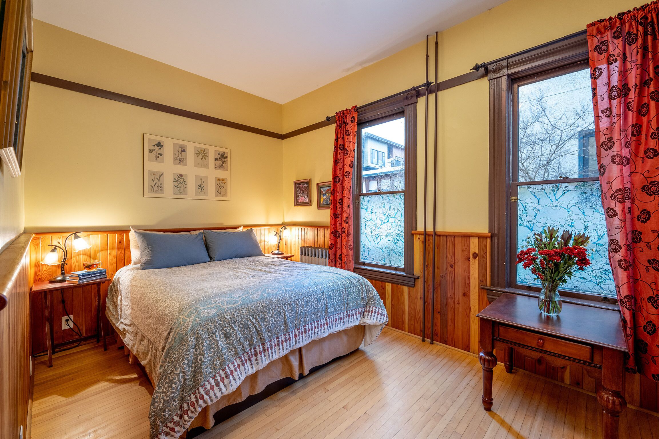 Um quarto aconchegante com uma cama grande, cabeceira de madeira, mesa de cabeceira com abajur, cômoda e uma janela com cortinas.