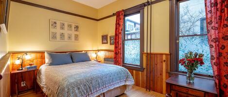 Um quarto aconchegante com uma cama grande, cabeceira de madeira, mesa de cabeceira com abajur, cômoda e uma janela com cortinas.