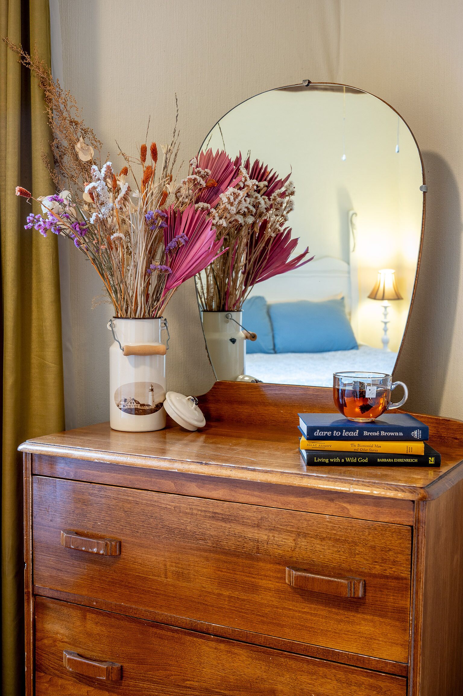 Een houten dressoir met een spiegel, een vaas met gedroogde bloemen, een glas thee en een stapel boeken.