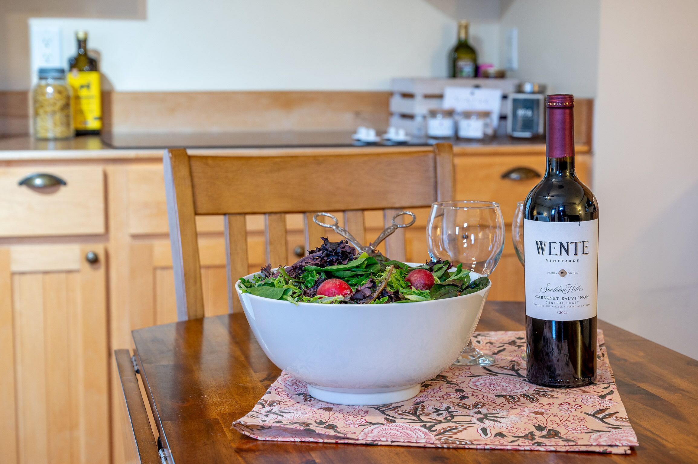 A wooden dining table with a white bowl of salad and a bottle of Wente wine.