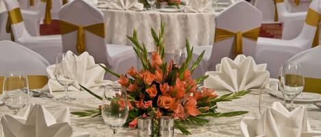 Uma sala de banquetes com mesas redondas preparadas para um evento formal, cada uma com toalhas brancas, fitas douradas e arranjos florais.