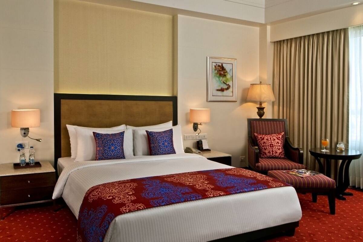 Ein Hotelzimmer mit einem großen Bett, zwei Nachttischen mit Lampen, einem Sessel, einem kleinen Tisch mit einem Getränk und einem Fenster mit Vorhängen.