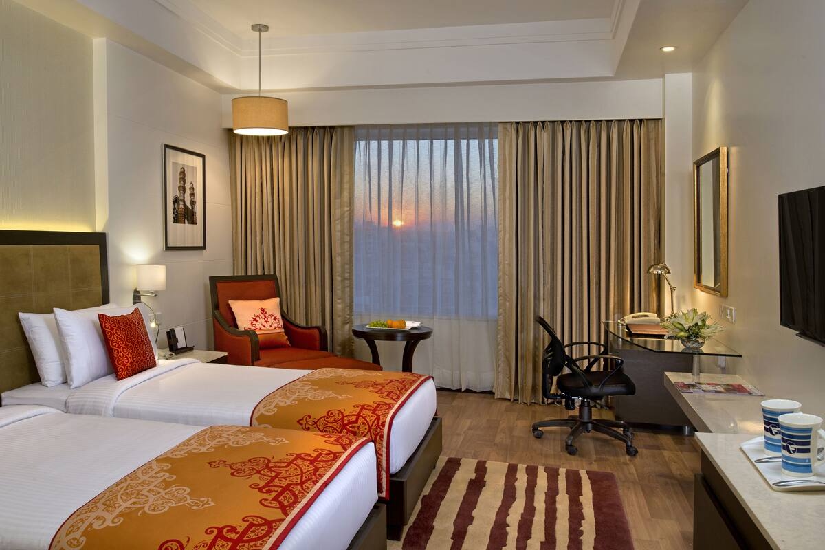 Ein Hotelzimmer mit zwei Betten, einem Schreibtisch, einem Stuhl, einem kleinen Tisch mit Früchten und Blick auf den Sonnenuntergang durch das Fenster.