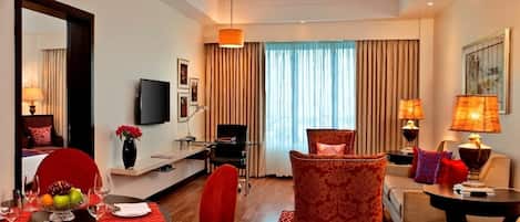 A hotel room with a dining area, a sofa, a TV, and a large window with curtains.