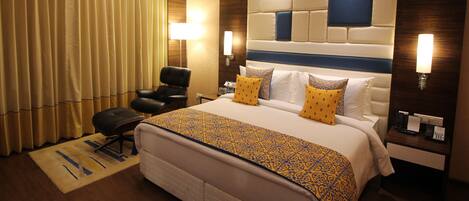 A hotel room with a large bed, a desk with a lamp, a chair, and a window with curtains.
