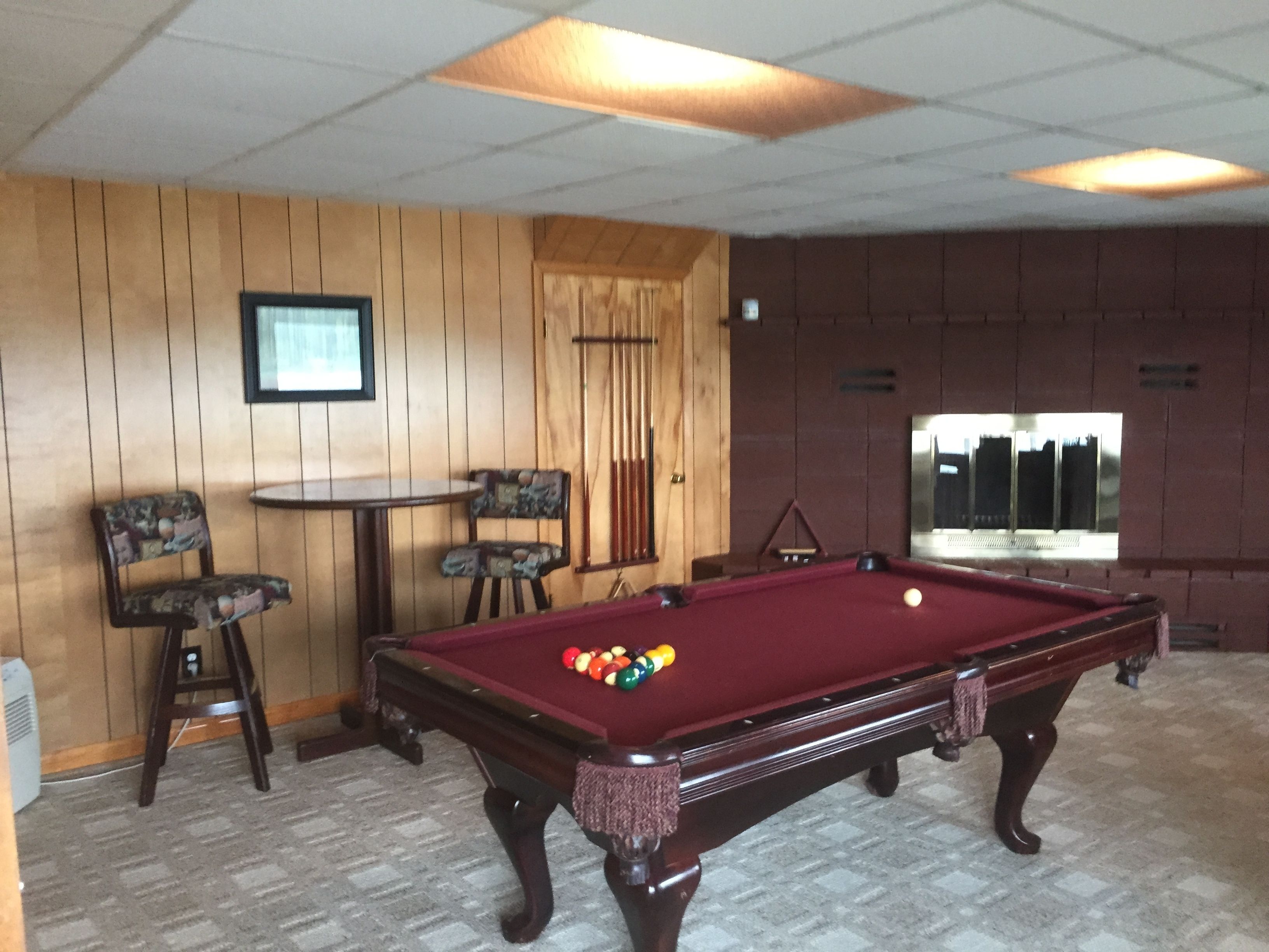 A pool table with balls, a pool cue, and two chairs in a room with wooden paneling and a window.