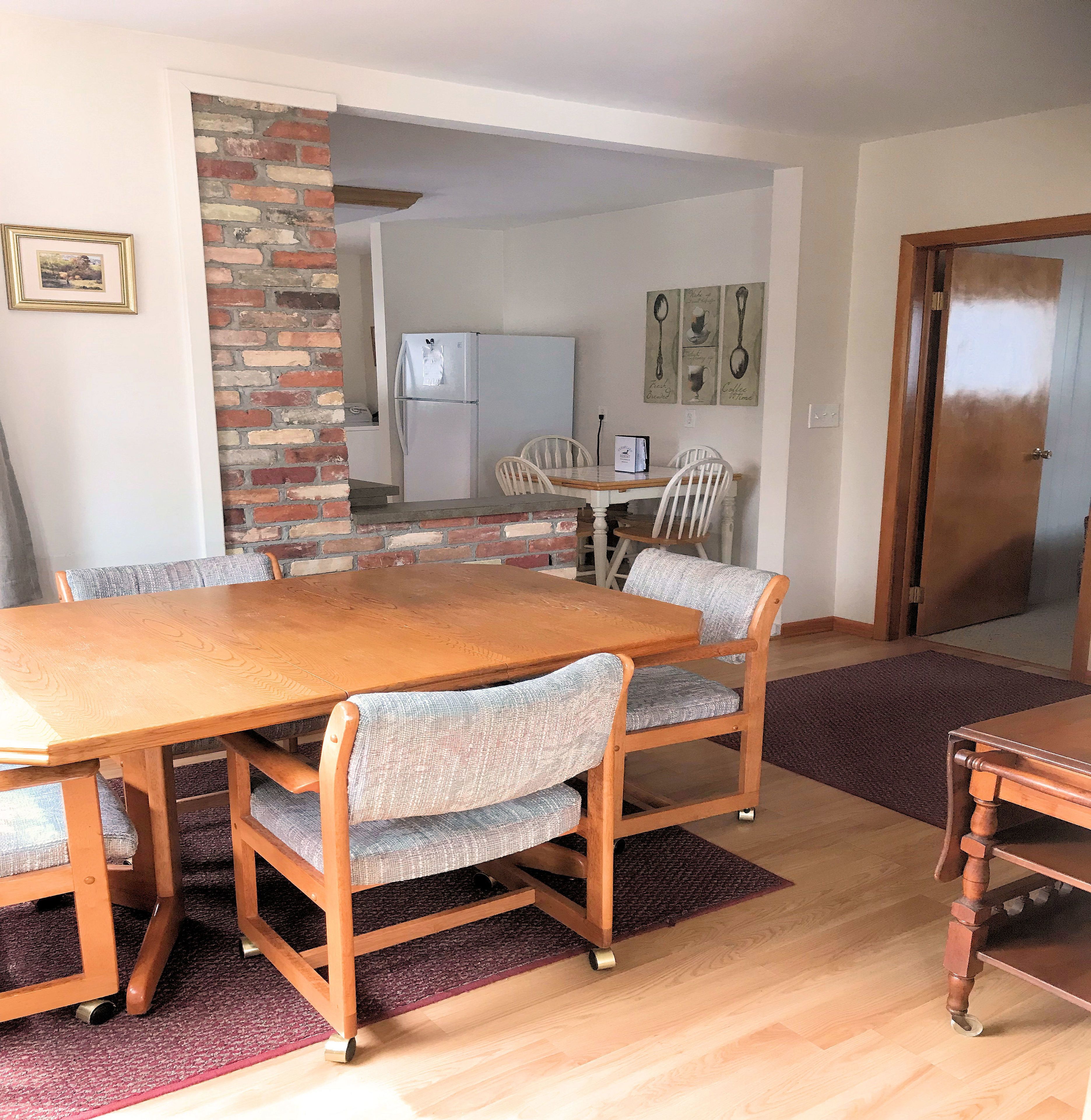 A dining area with a wooden table and chairs, a brick column, a refrigerator, and a door.