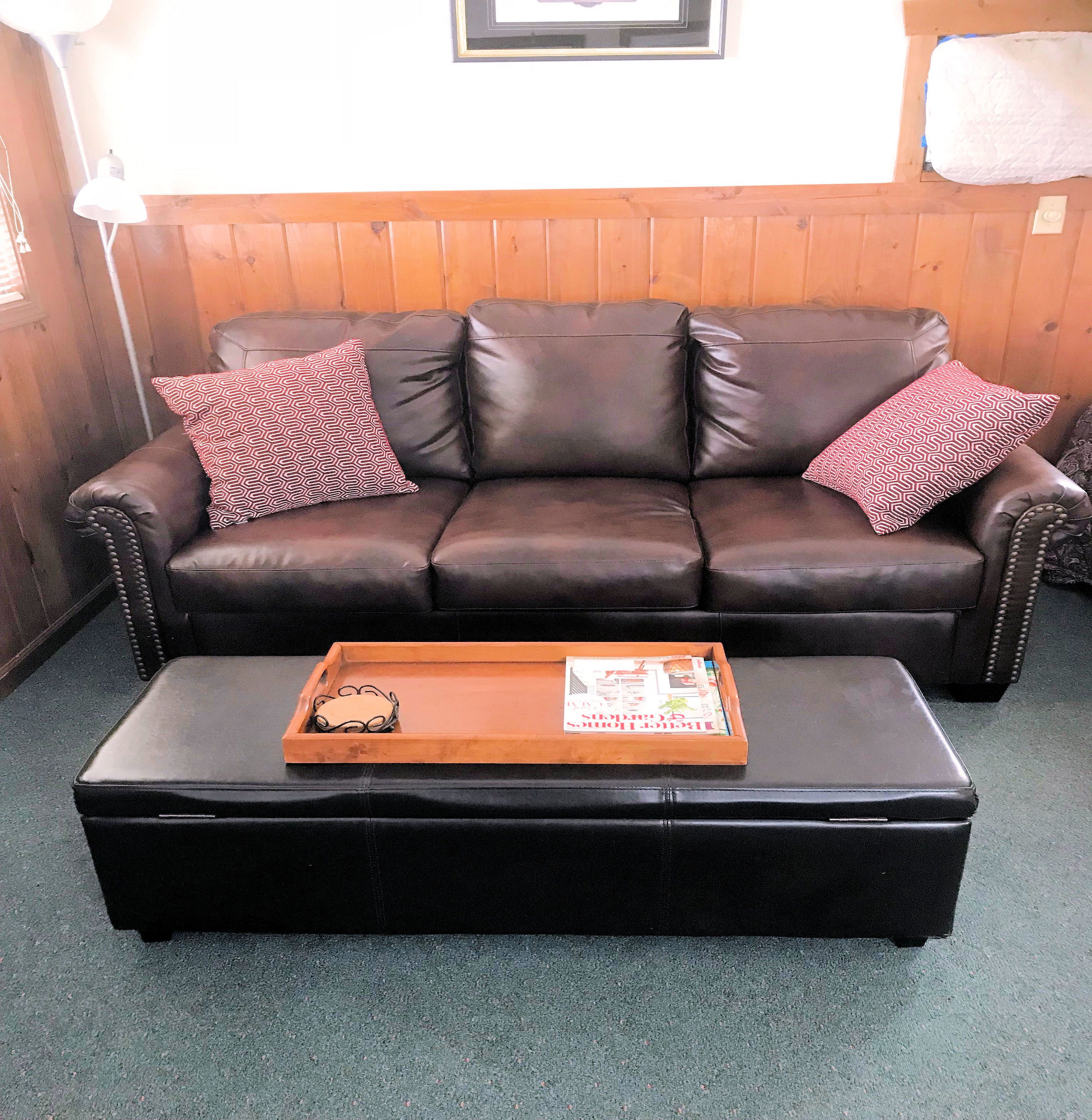 A brown leather sofa with patterned pillows, a wooden tray with magazines, and a black ottoman.