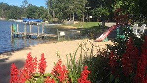 Un parque junto al lago con un muelle de madera, un área de juegos y flores rojas en primer plano.