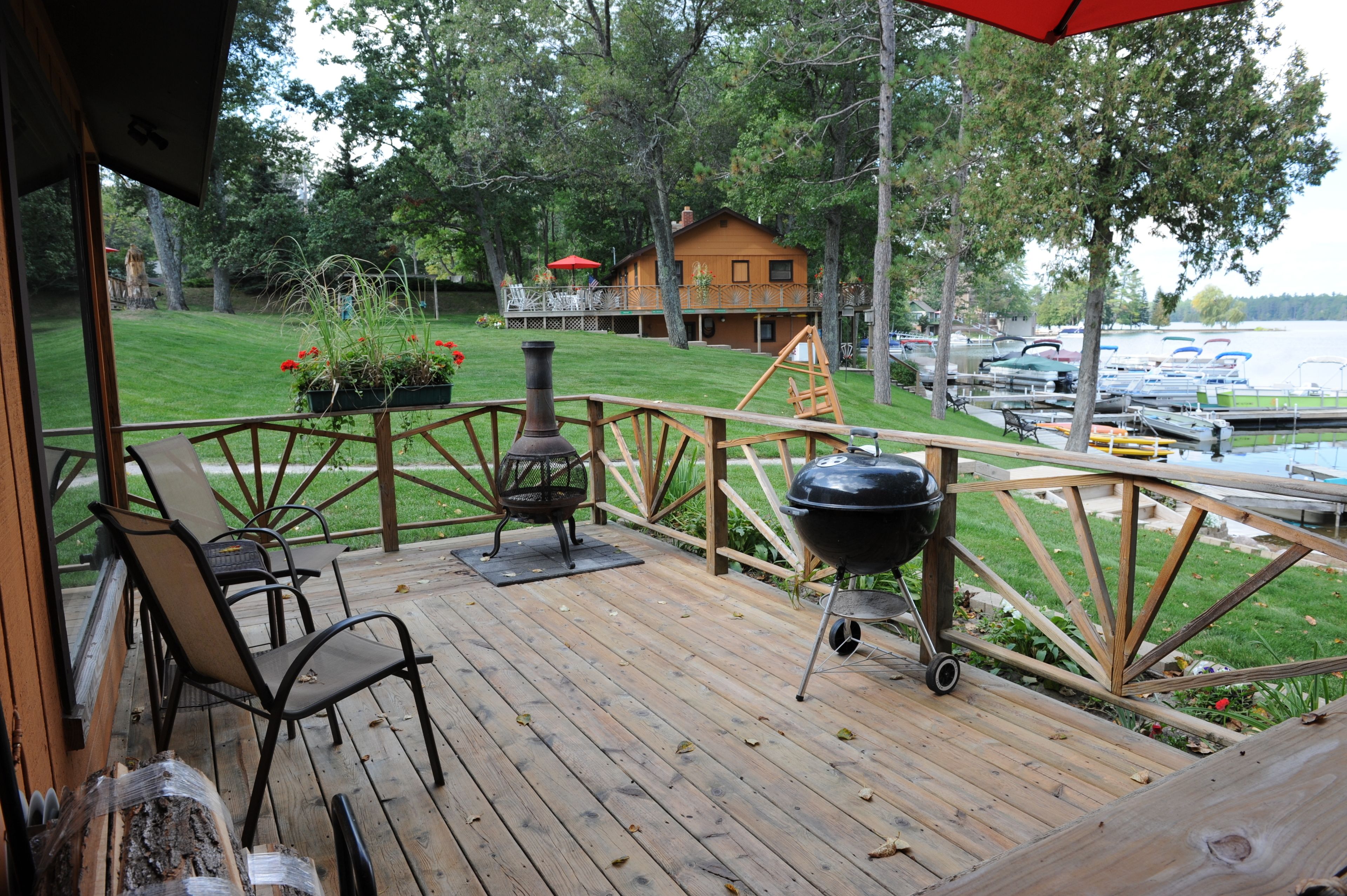 Een houten terras met buitenmeubilair, een barbecue en uitzicht op een meer met boten.