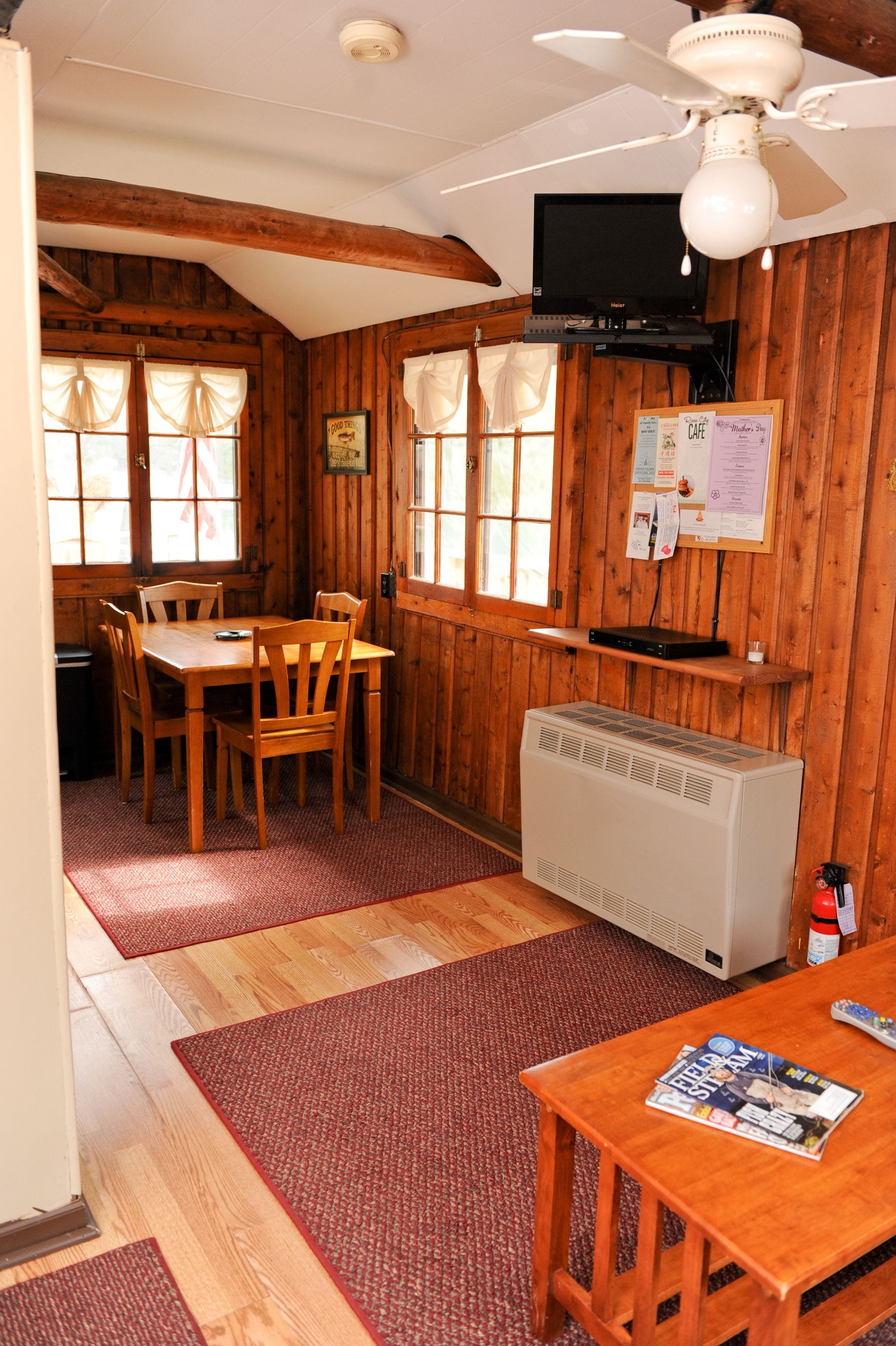 A wooden cabin interior with a dining table, chairs, a ceiling fan, a TV mounted on the wall, and a fireplace.