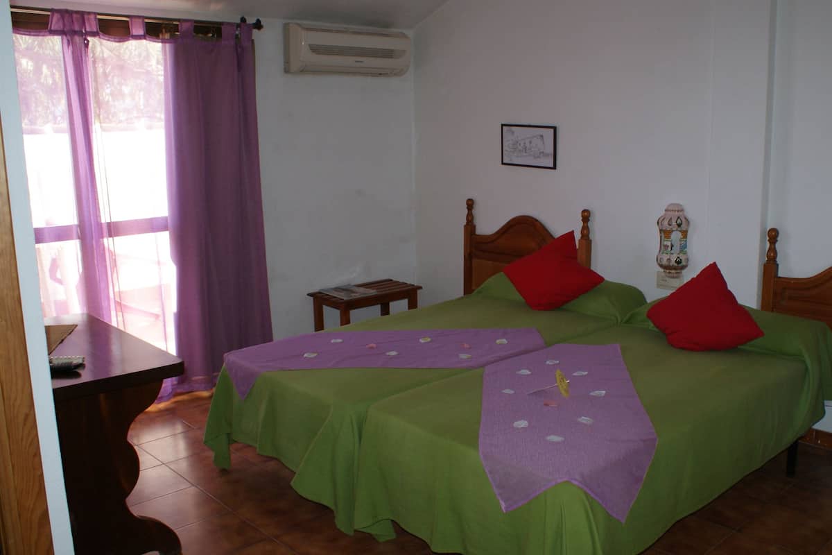 Una habitación con dos camas, un escritorio de madera, una mesita pequeña y un ventanal con cortinas moradas.