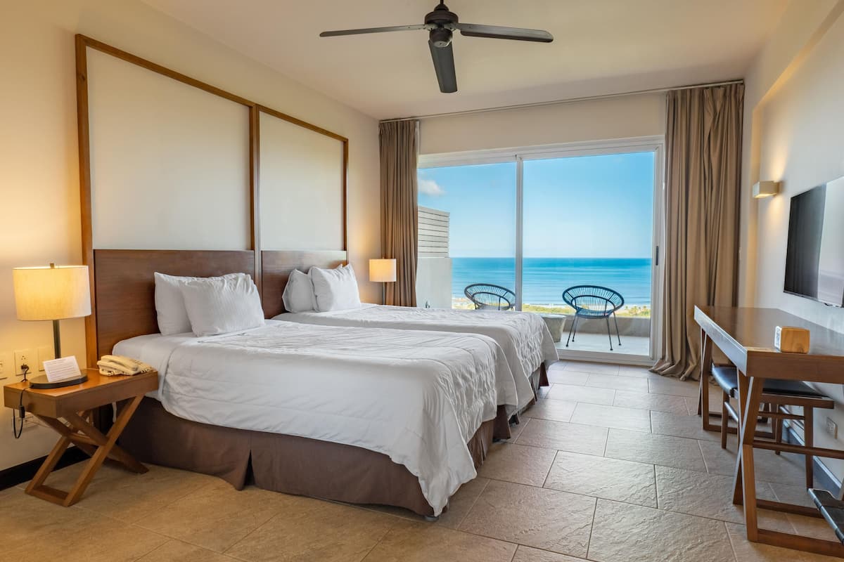A hotel room with two beds, a desk, a TV, and a balcony with a view of the ocean.