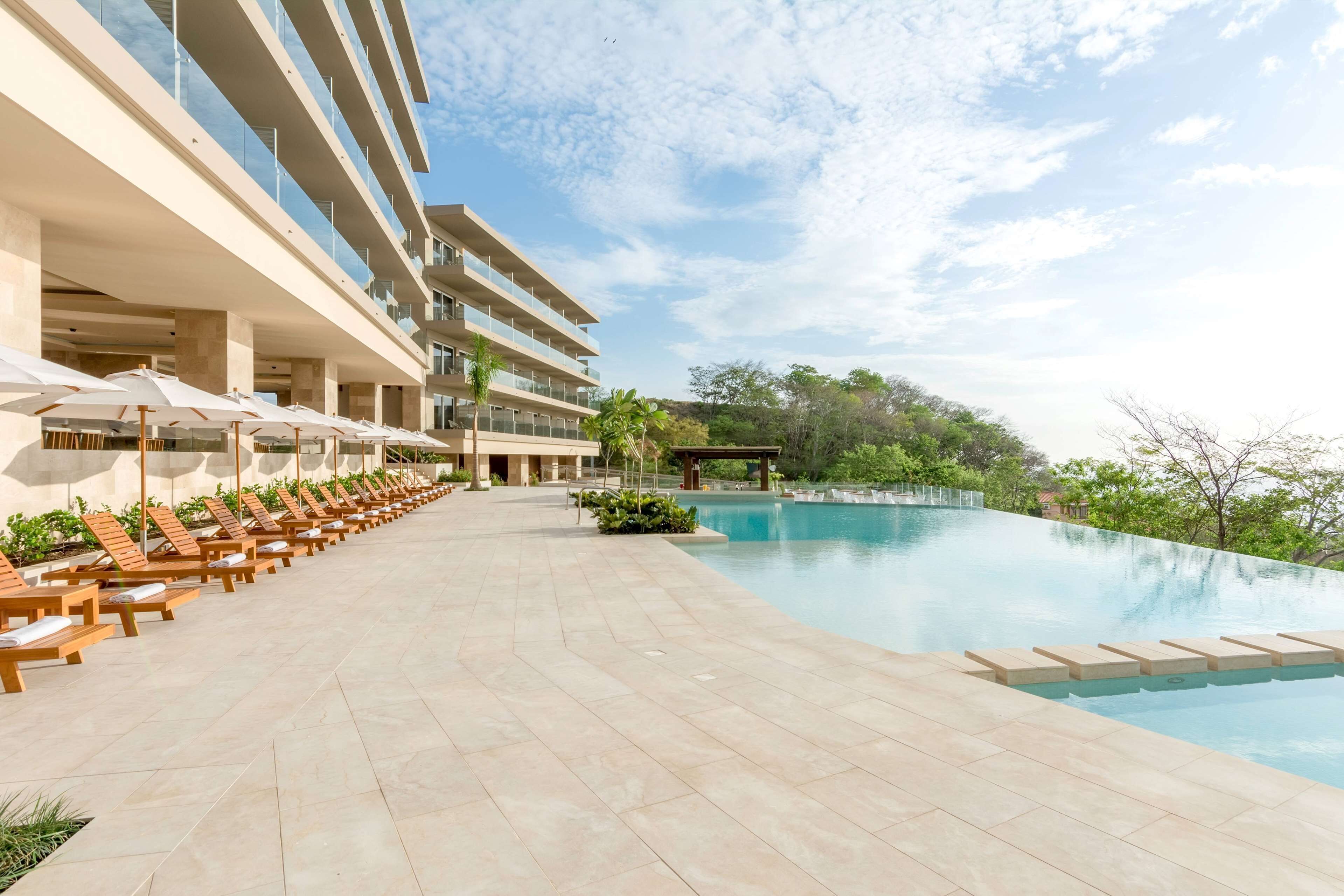 A hotel pool area with sun loungers and umbrellas, a clear sky, and a large pool.