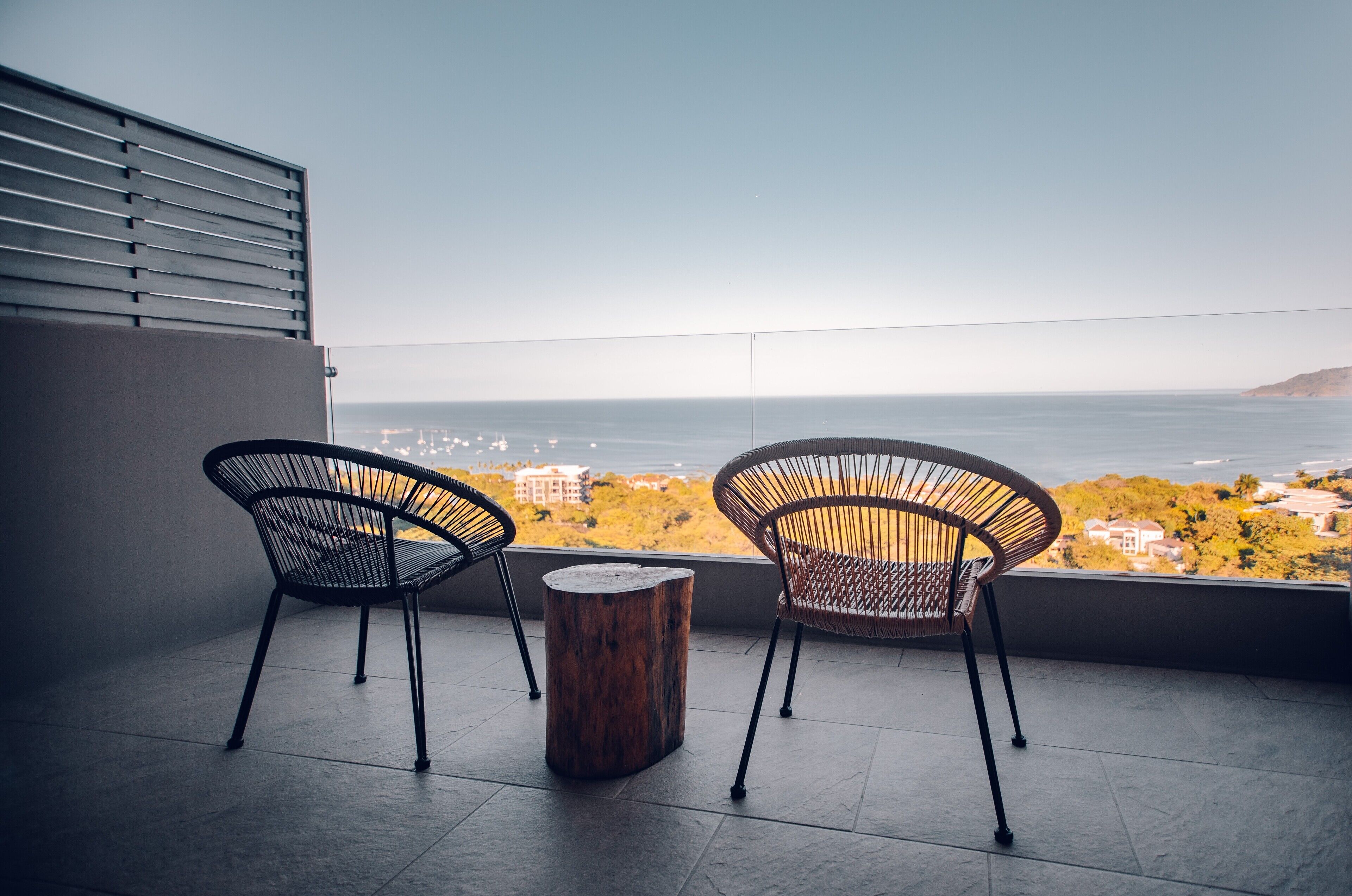 Zwei Korbstühle und ein Holzschemel auf einem Balkon mit Blick auf das Meer und Gebäude.