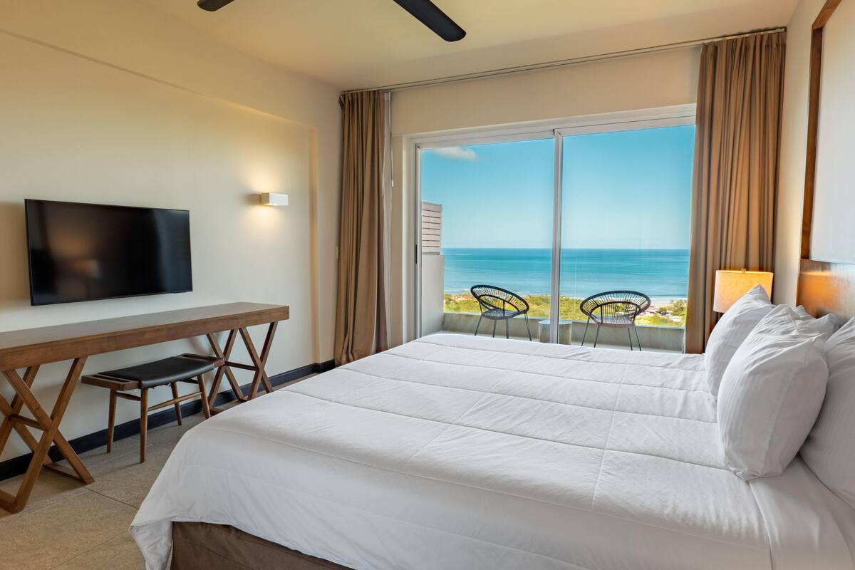 A hotel room with a large bed, a TV, a desk with a chair, and a view of the ocean.