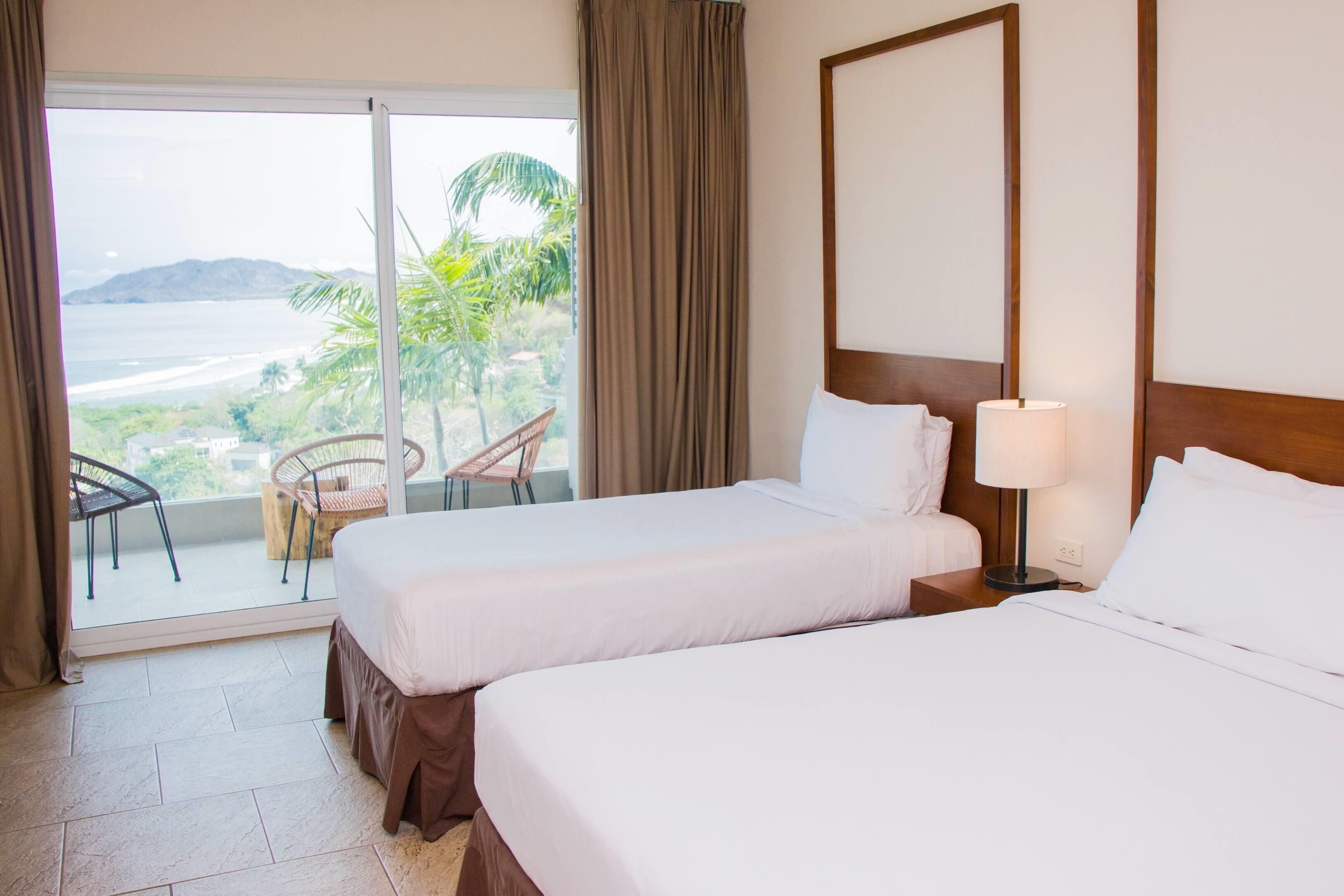 A hotel room with two beds, a balcony with a view of the ocean, and a chair.