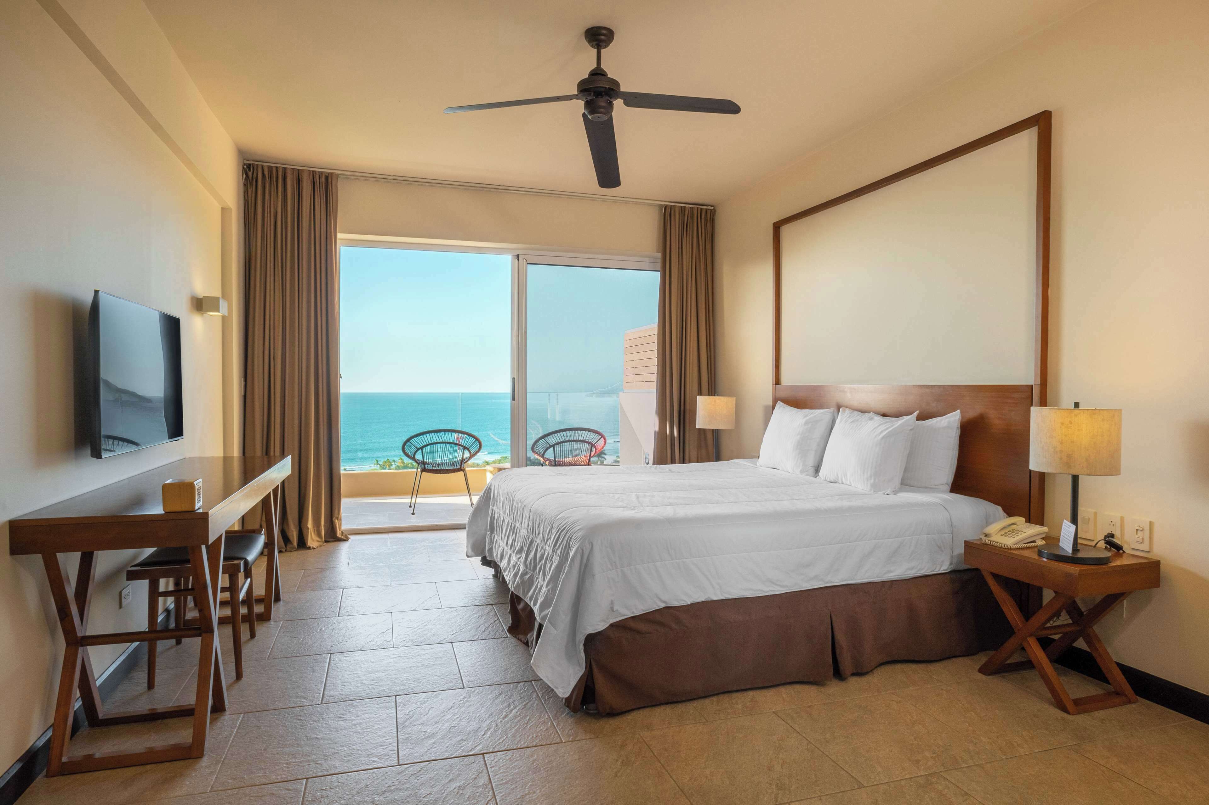A hotel room with a bed, a desk, a chair, a TV, a ceiling fan, and a view of the ocean.