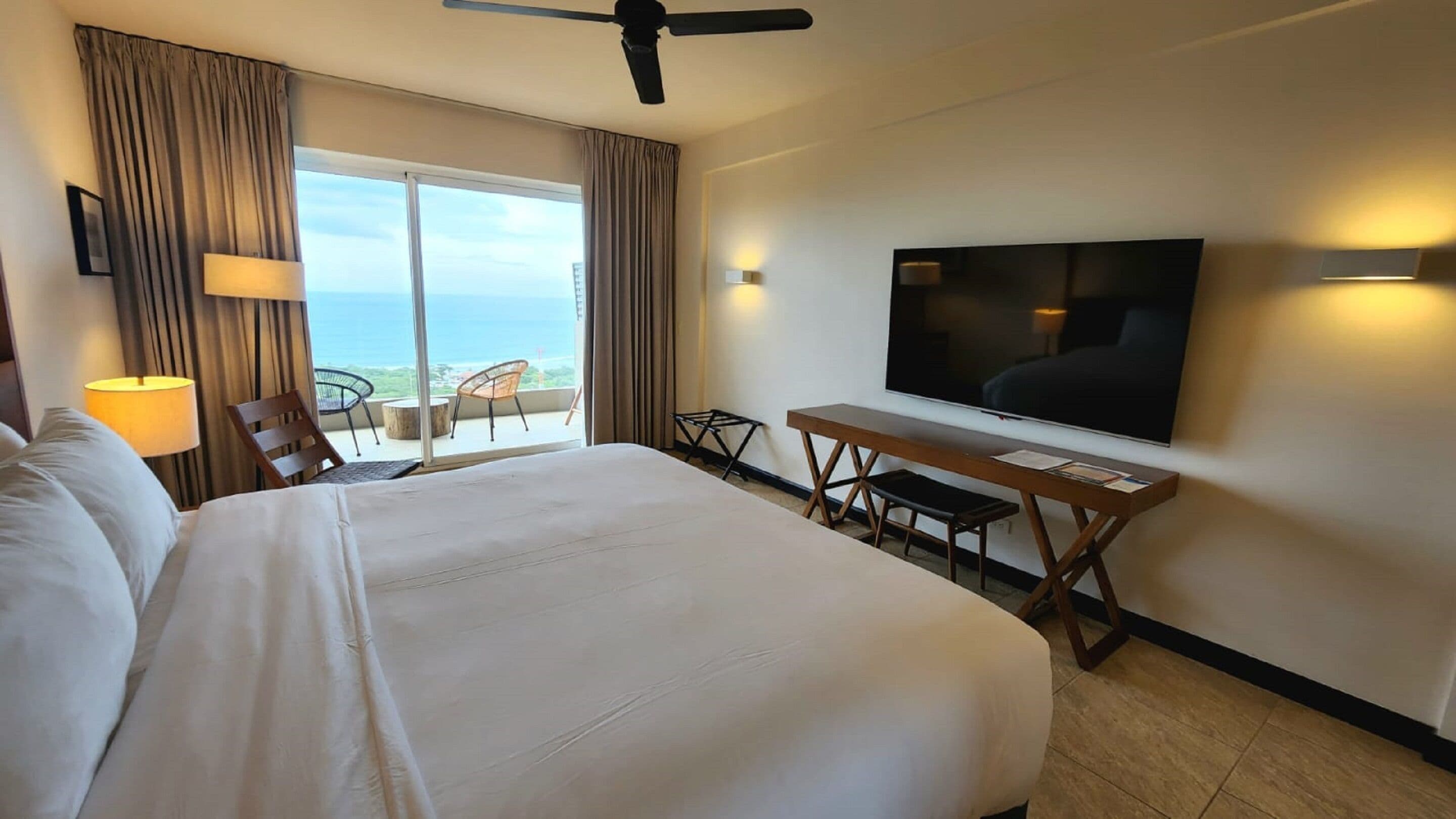 A hotel room with a large bed, a desk with a flat-screen TV, a chair, and a balcony with a view of the ocean.