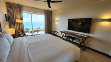 A hotel room with a large bed, a desk with a flat-screen TV, a chair, and a balcony with a view of the ocean.