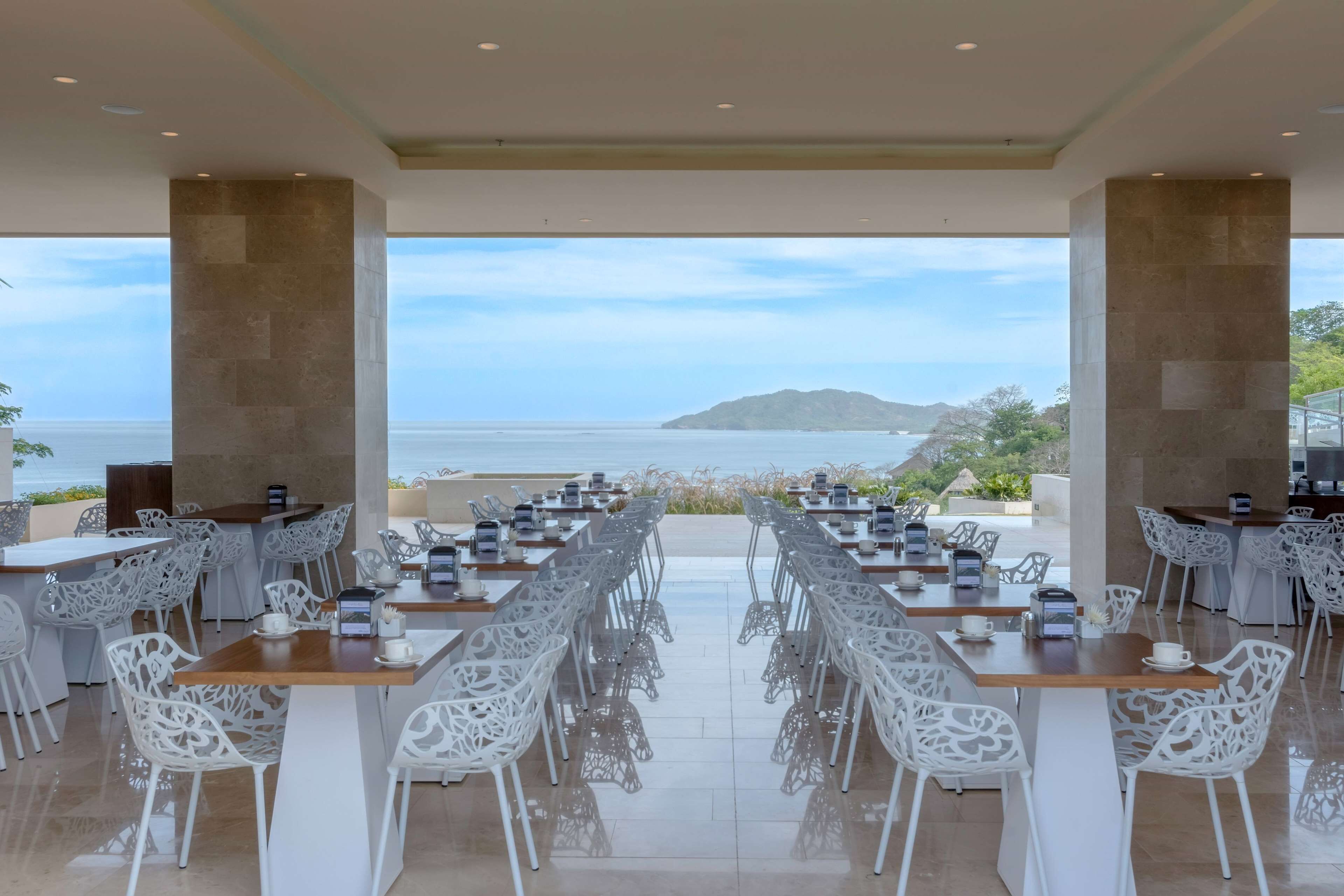 Comedor con sillas y mesas blancas, ventanas amplias con vista al mar y una isla distante.