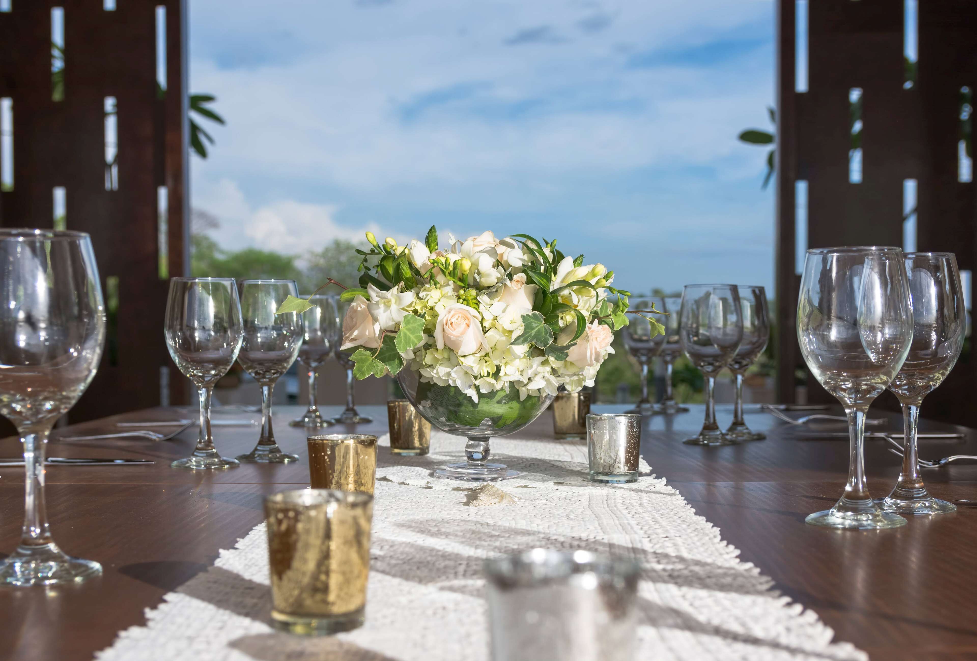 A table set with wine glasses and a centerpiece of flowers, with a view of the sky and greenery in the background.