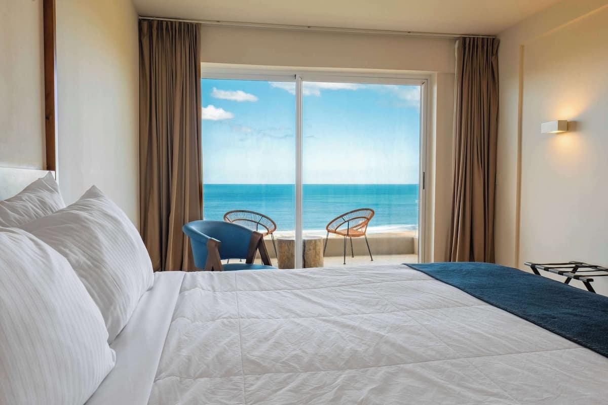 A hotel room with a large bed, two chairs, a view of the ocean, and a sliding glass door.