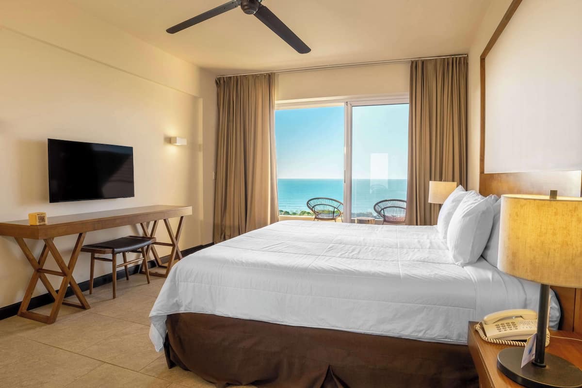 A hotel room with a bed, a desk, a television, a phone, and a view of the ocean.
