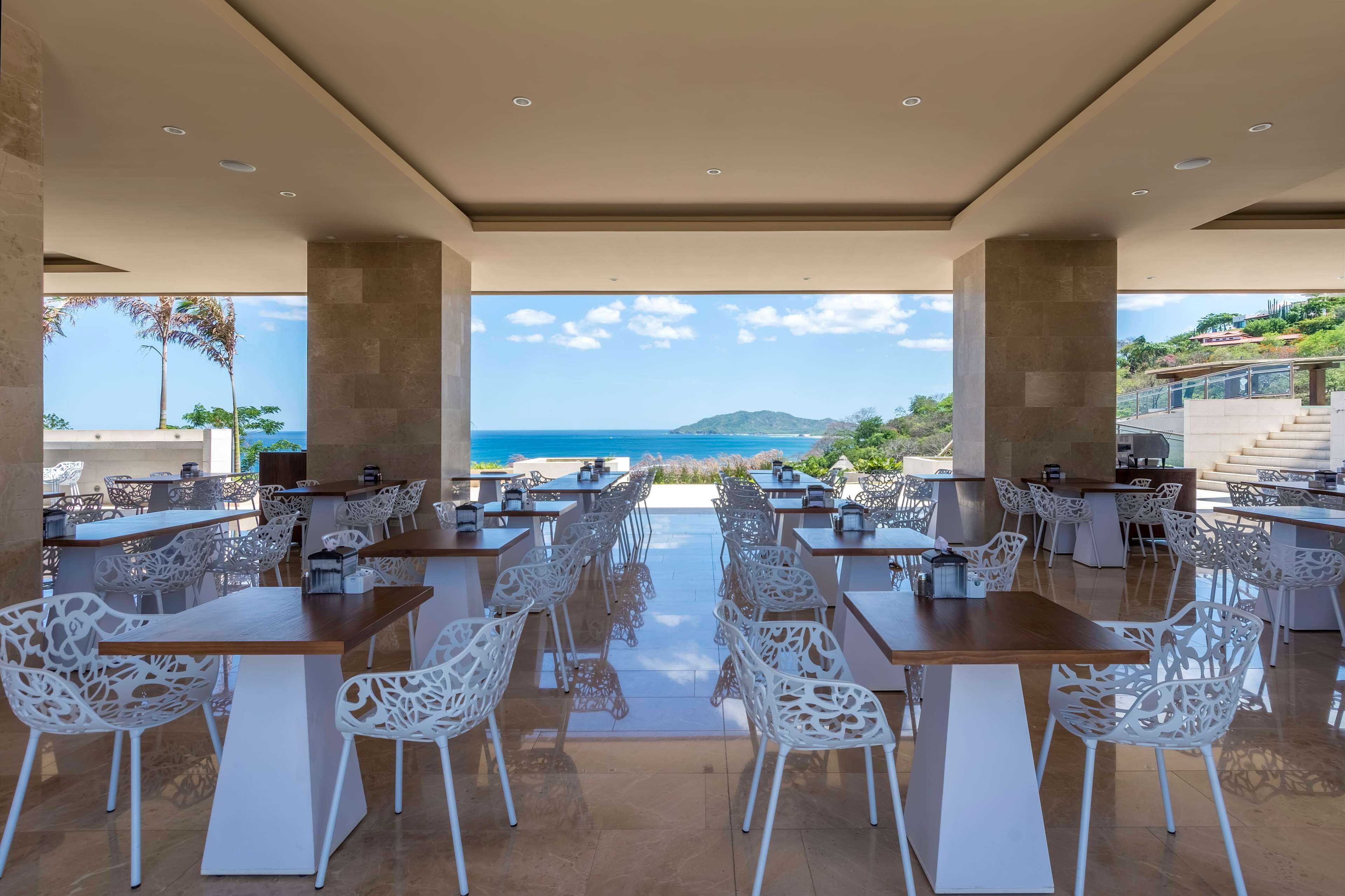 Un área de comedor al aire libre con mesas y sillas, vista al mar y un paisaje montañoso de fondo.