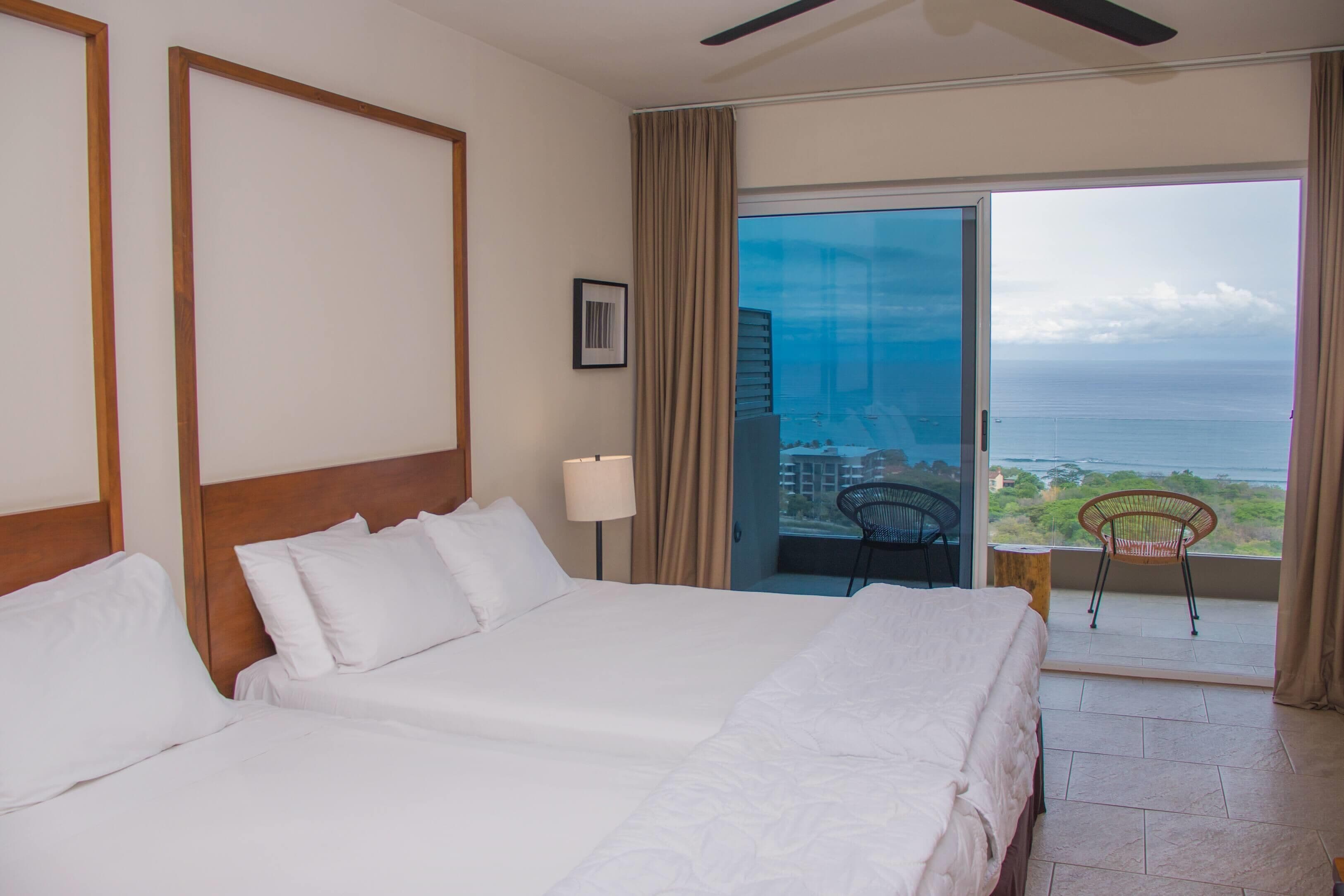 Habitación de hotel con una cama grande, ventilador de techo y balcón con vista al océano.