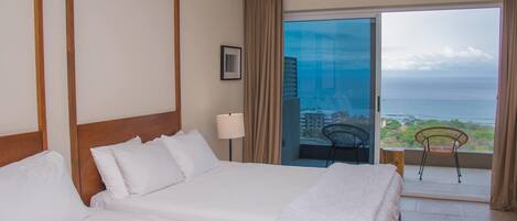 A hotel room with a large bed, a ceiling fan, and a balcony with a view of the ocean.