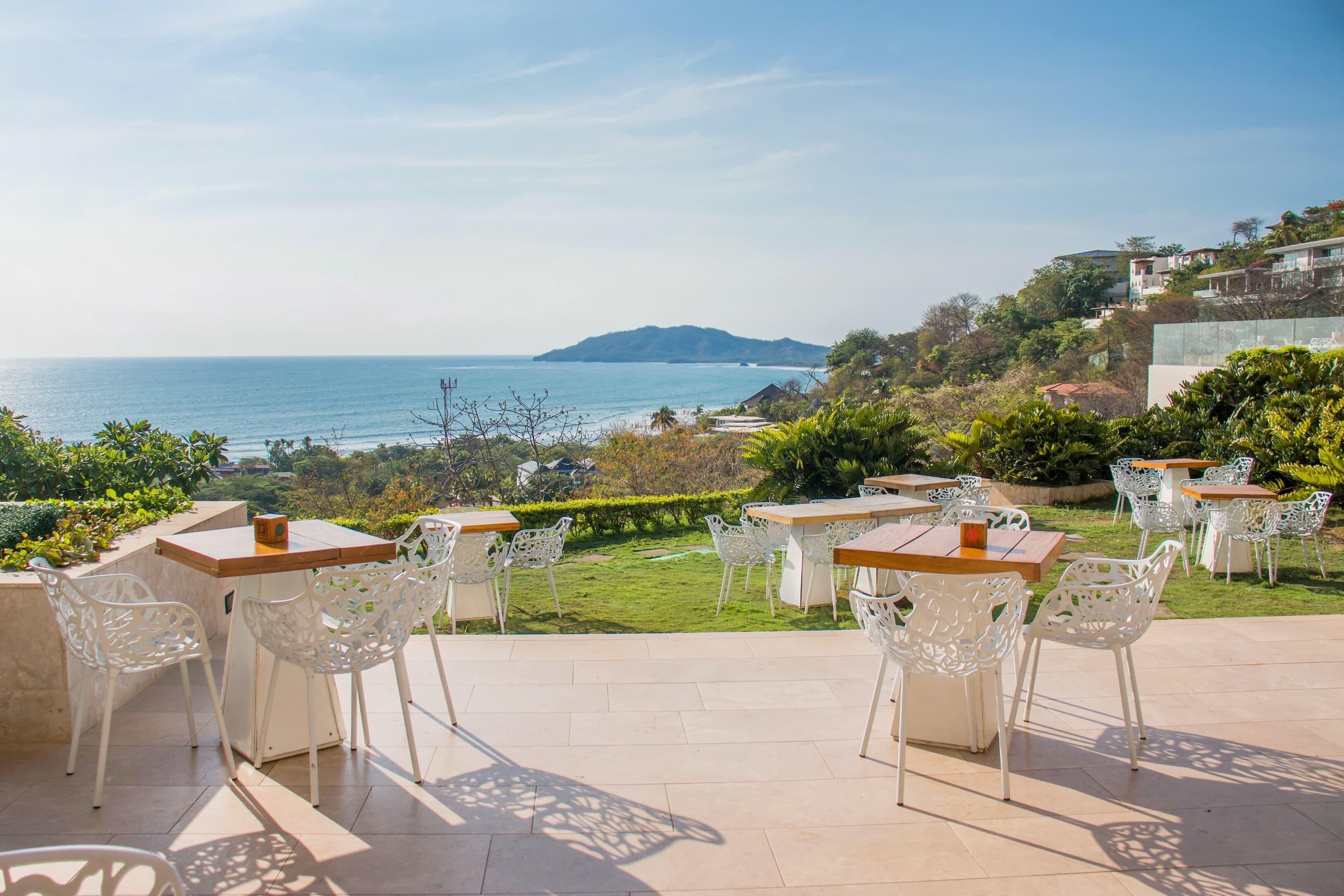 Un espace extérieur aménagé avec des chaises et des tables blanches, offrant une vue sur la côte, avec une montagne lointaine et une végétation luxuriante.