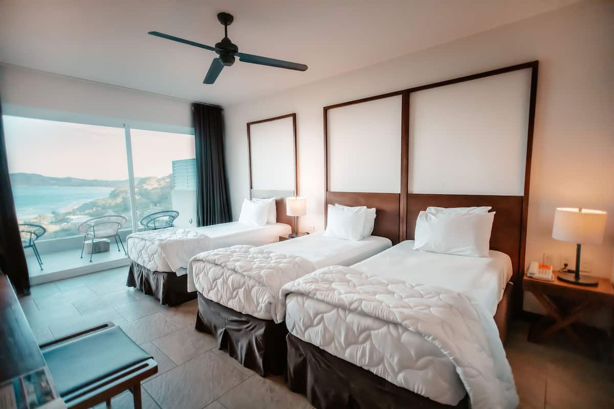 A hotel room with two beds, a ceiling fan, a balcony with a view of the ocean, and a desk with a chair.