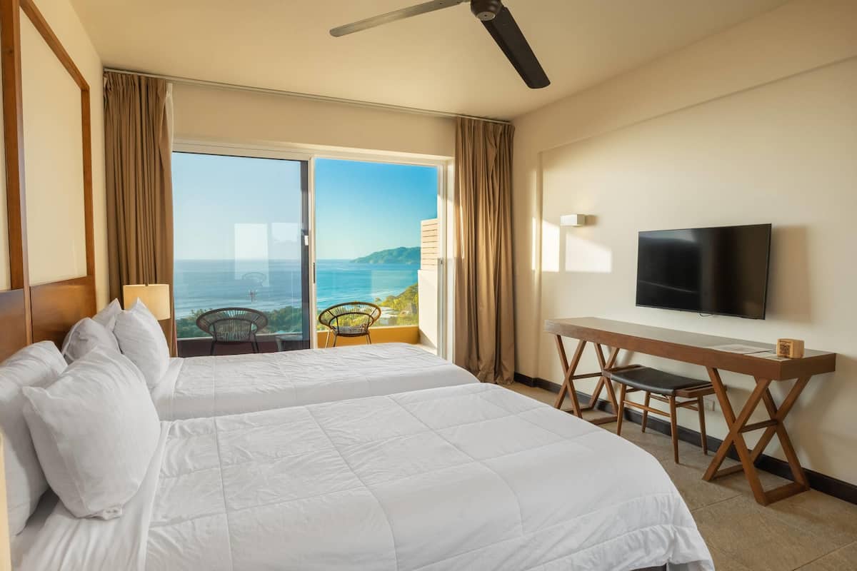 A hotel room with two beds, a desk, a chair, a TV, and a balcony with a view of the sea.