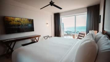 A hotel room with a bed, a desk, a chair, a ceiling fan, and a large window with a view of the sea.