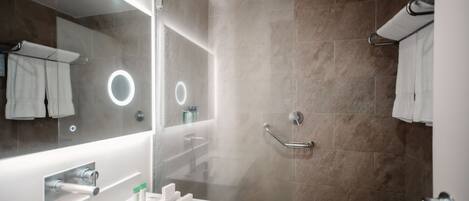 Un baño moderno con ducha envuelta en vidrio, un espejo grande con iluminación integrada y un tocador blanco con artículos de aseo.
