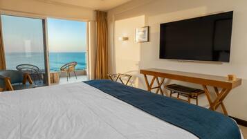 A hotel room with a large bed, a desk, and a TV. There is a balcony with a view of the ocean.