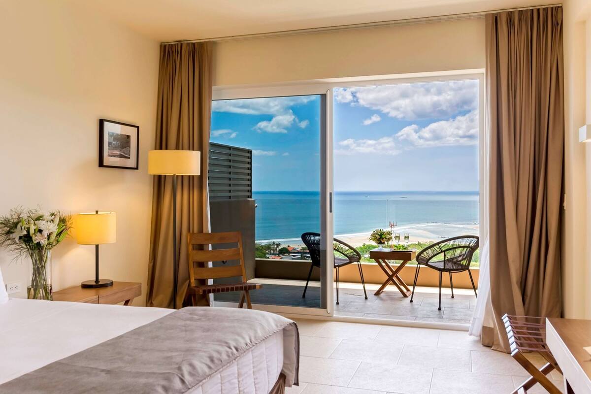 A hotel room with a bed, a nightstand with a lamp, a chair, and a balcony with a view of the ocean.