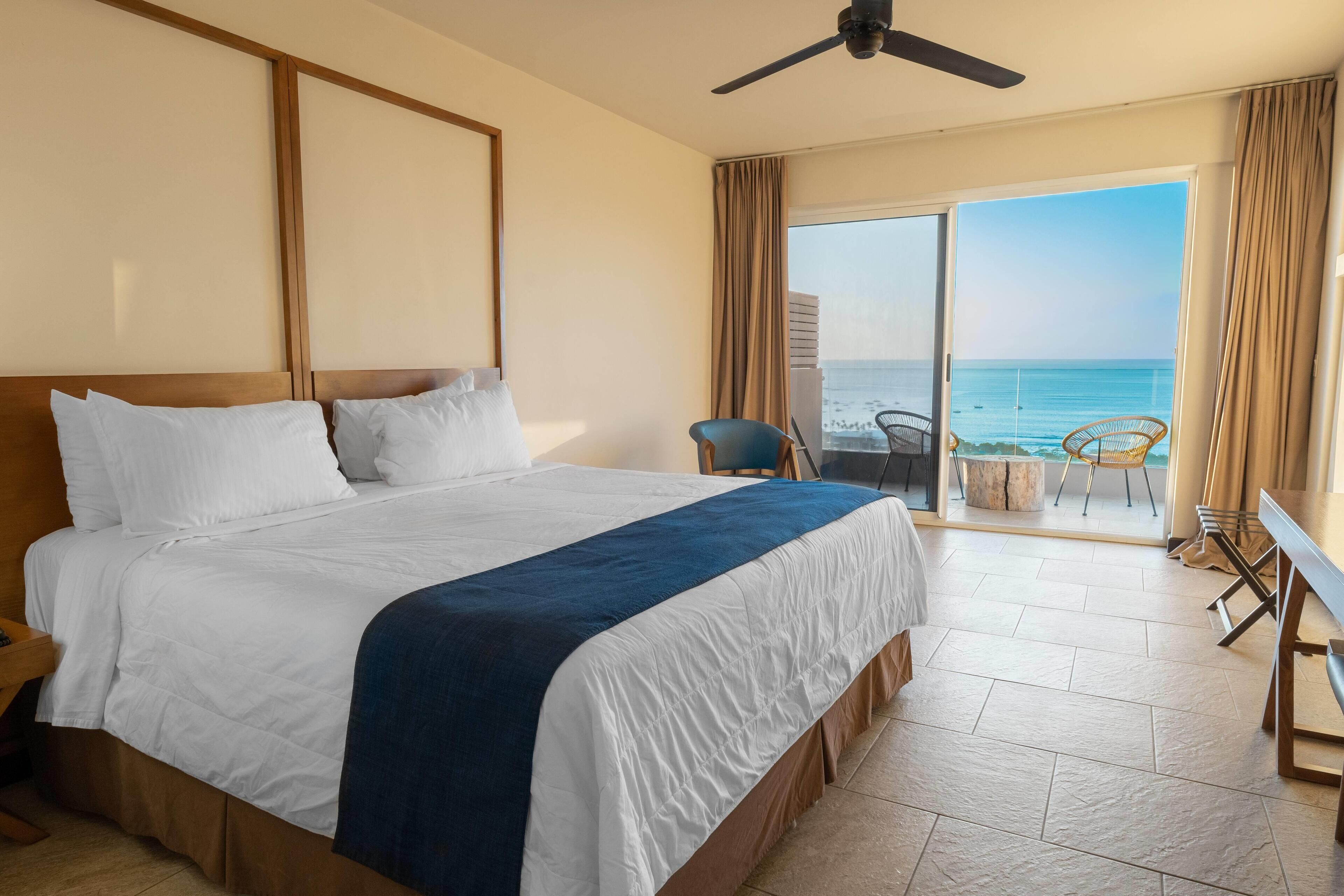 A hotel room with a large bed, a ceiling fan, a balcony with a view of the ocean, and a seating area with chairs.