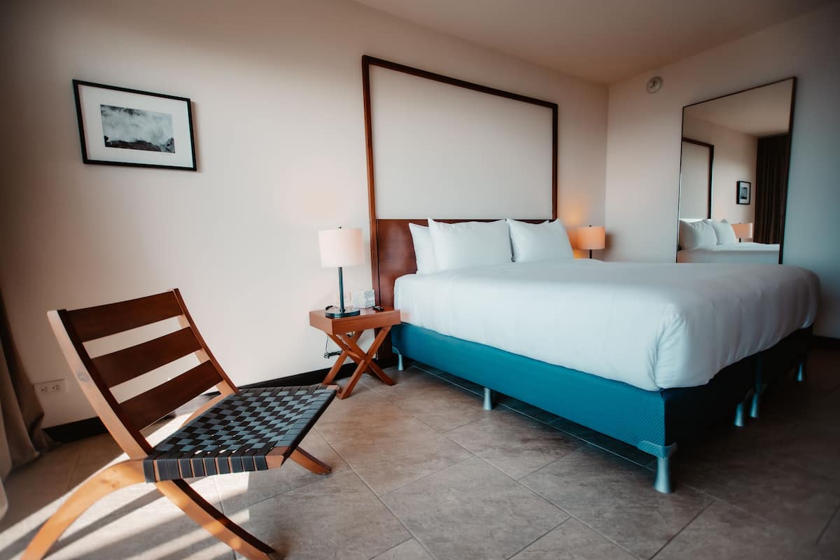 A hotel room with a bed, a chair, a small table, a lamp, and a framed picture on the wall.