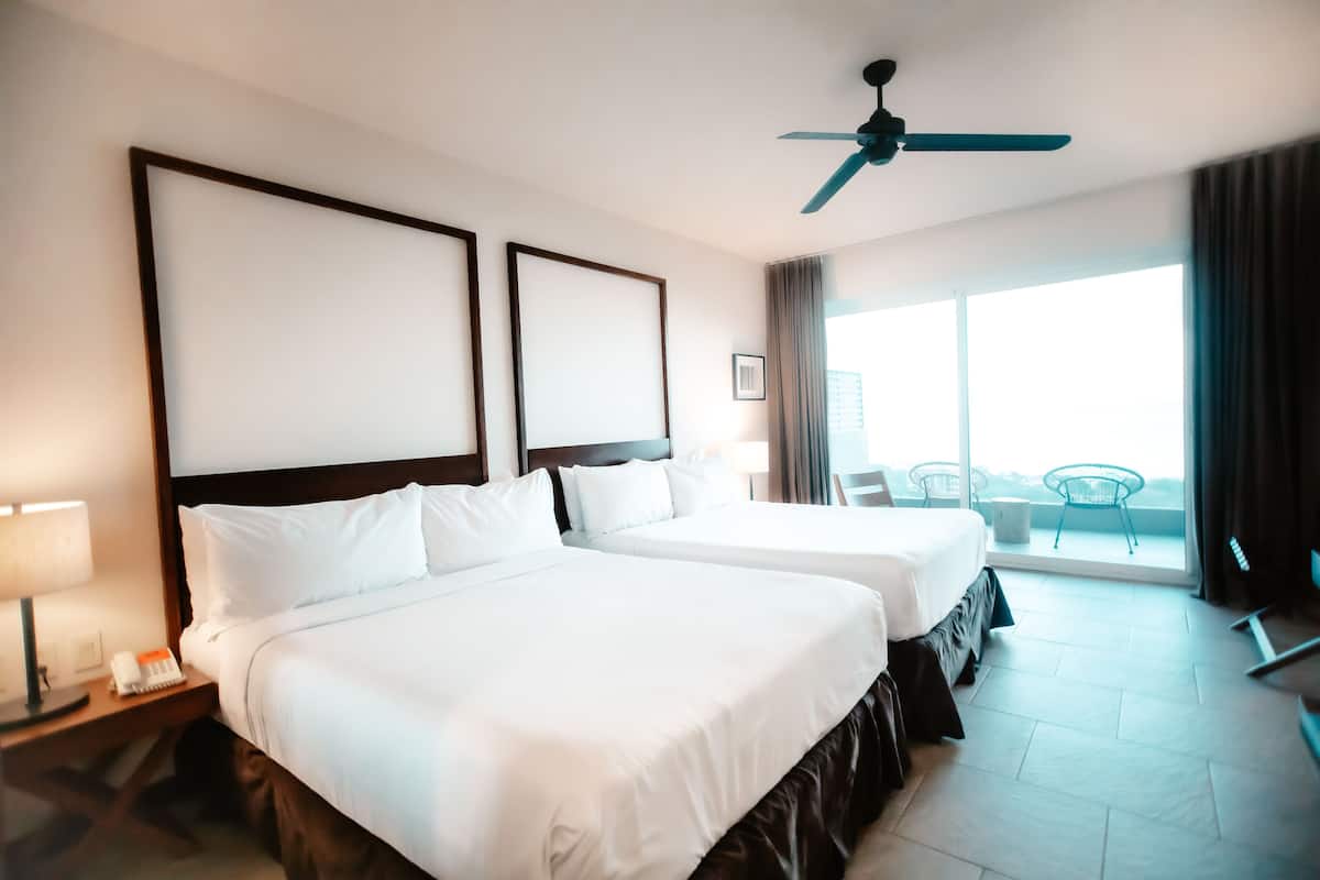 A hotel room with two beds, a ceiling fan, a balcony with a view, and a nightstand with a lamp and phone.