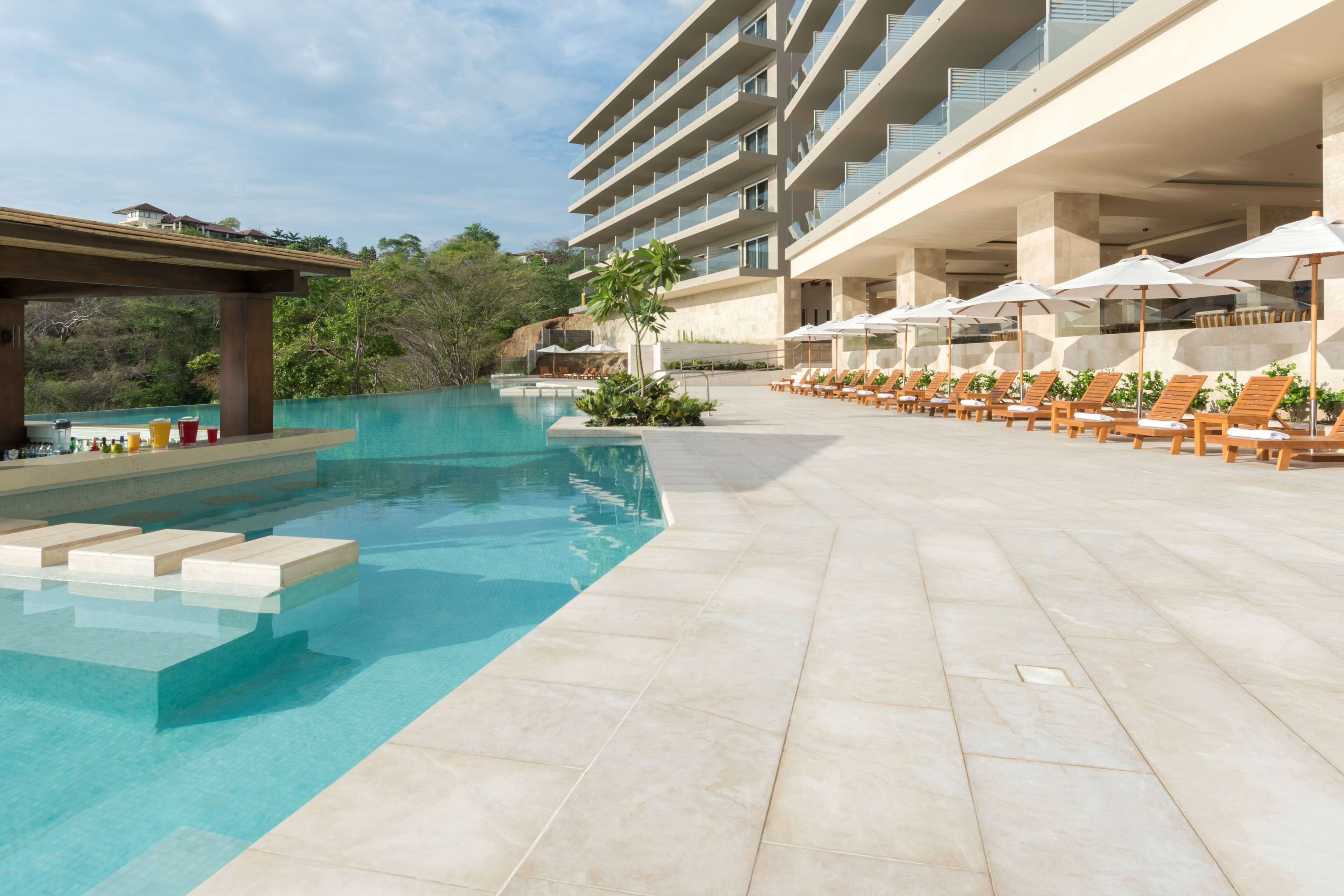 A pool area with lounge chairs and umbrellas, a covered seating area, and a building with multiple balconies.