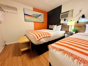 A hotel room with two beds, a wooden floor, a wall-mounted air conditioner, a nightstand with a lamp, and a painting.
