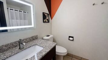 A bathroom with a granite countertop sink, a white toilet, a mirror with a light, and a framed picture on the wall.