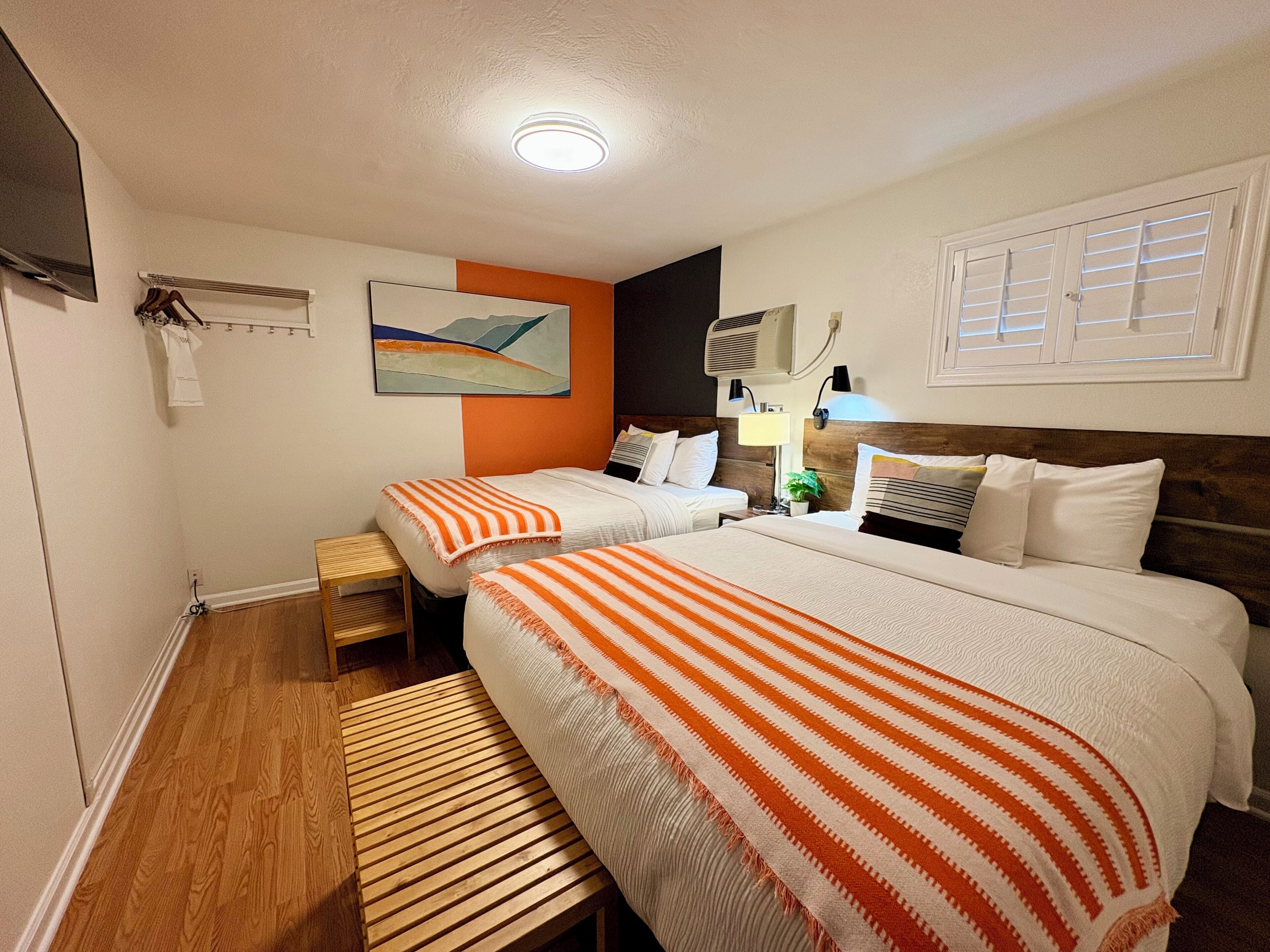 A hotel room with two beds, a wooden bench, a wall-mounted TV, and a ceiling light.