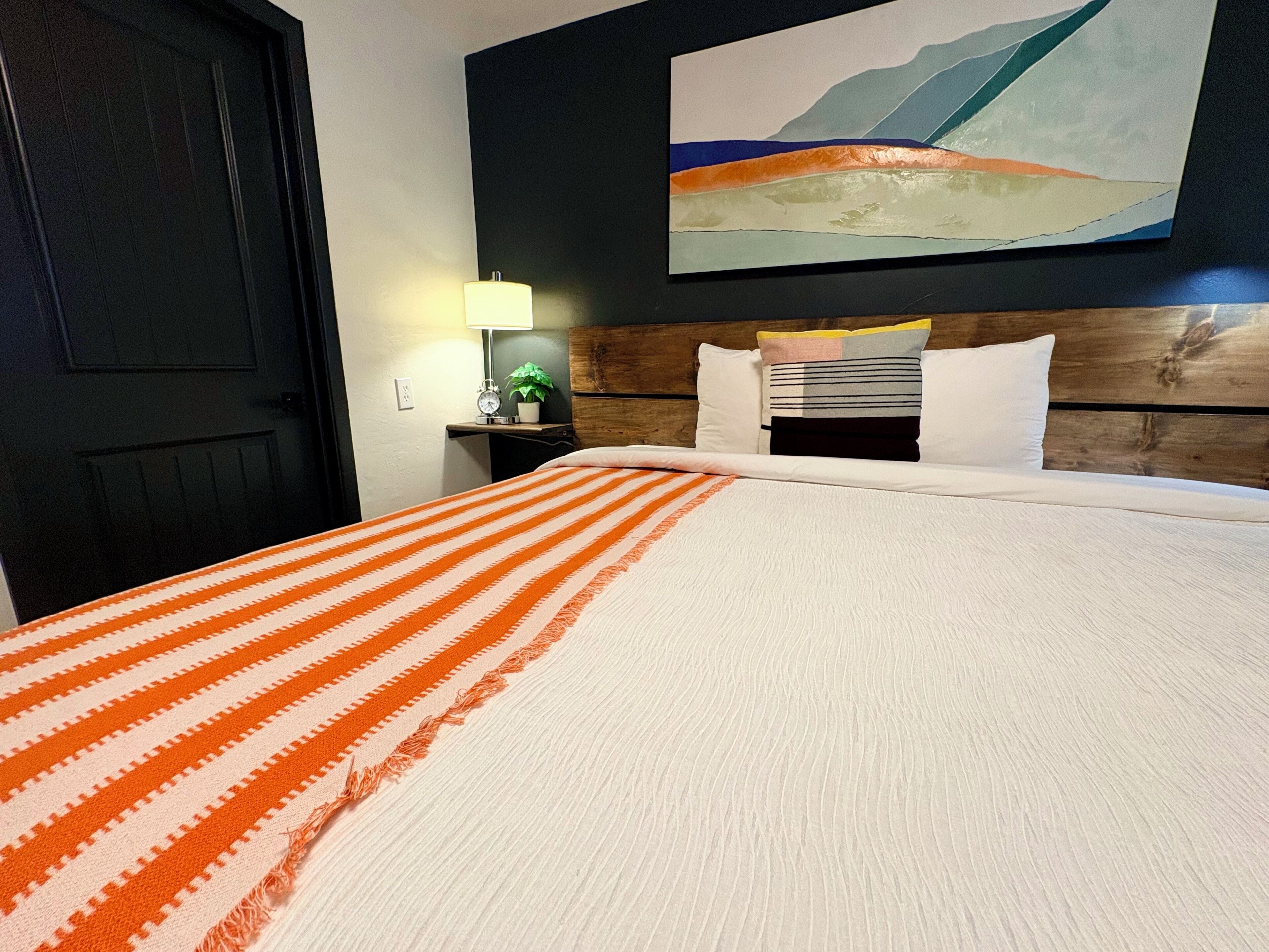 A hotel room with a bed, a nightstand with a lamp, a clock, and a wall painting.