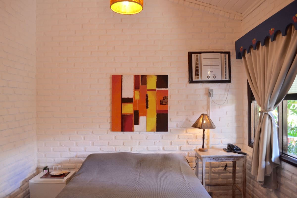A hotel room with a white brick wall, a bed, a bedside table with a lamp, a telephone, and a window with curtains.