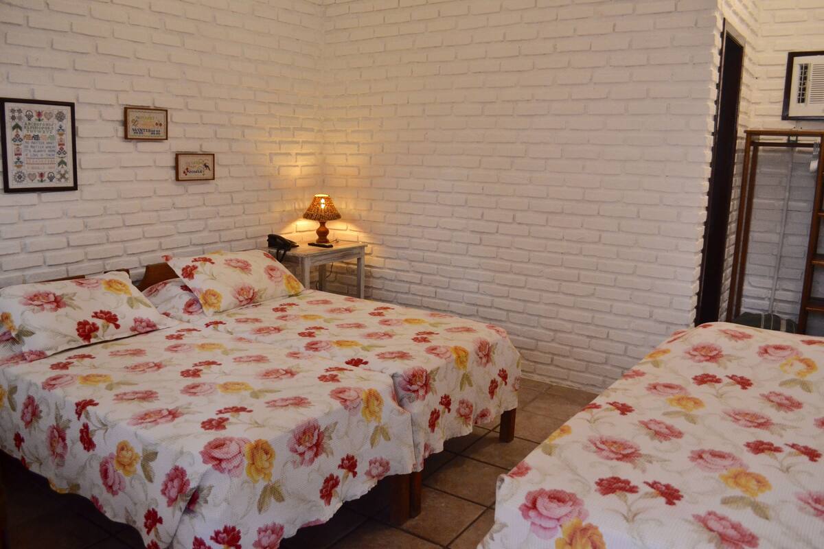 A room with two beds, a small table with a lamp, and a white brick wall.
